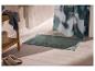 Tapis de bain vert avec bordure texturée sur carrelage beige, à côté d'une étagère en bois.