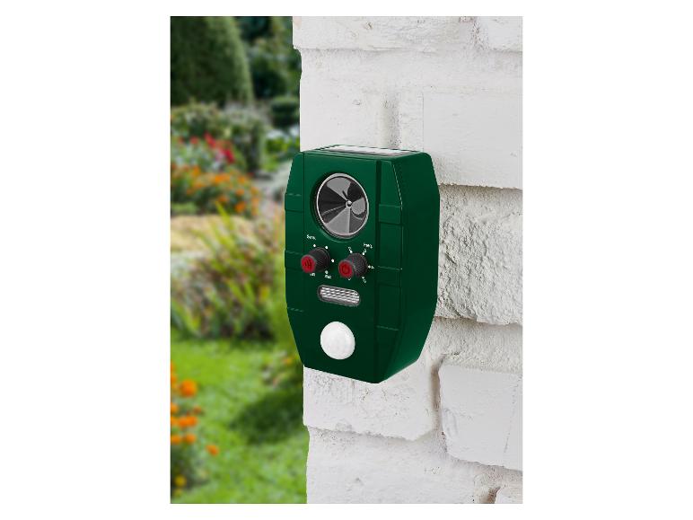 Répulsif animal vert à panneau solaire monté sur un mur de briques blanches dans un jardin.