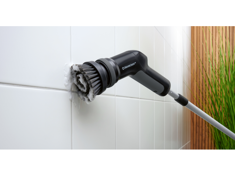 Brosse de nettoyage électrique pour carrelage avec mousse, devant un mur en bois et des plantes.