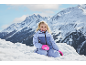 Fille souriante en combinaison de ski et gants roses dans la neige, avec des montagnes en arrière-plan.
