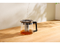 Théière en verre avec infuseur, où l'eau est versée, sur une table en bois.