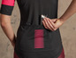 Une femme portant un maillot de vélo noir avec des détails rouges, rangeant une barre énergétique 'High Power' dans sa poche arrière.