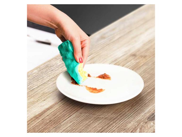 Main nettoyant une assiette avec une éponge à vaisselle sur une table en bois.