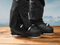 Des chaussures de ski noires sur un sol en bois avec des montagnes en arrière-plan.