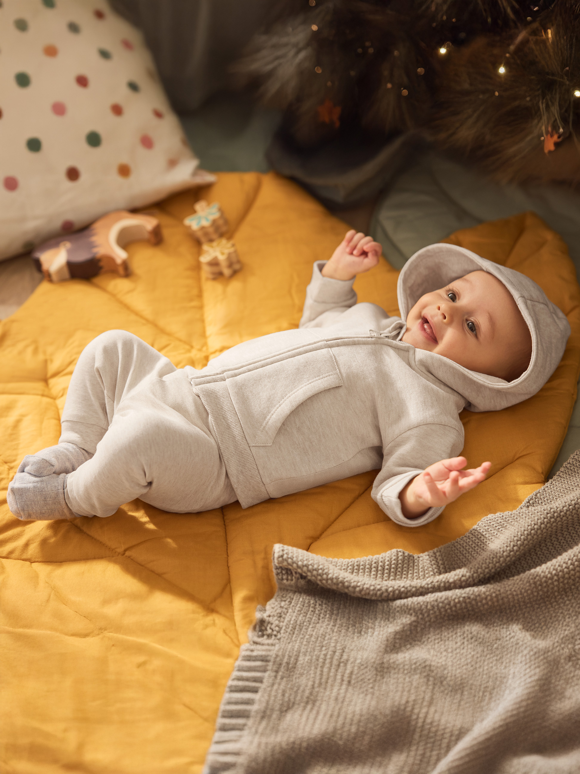 Bébé heureux en grenouillère grise sur matelas jaune avec jouets et lumières de Noël.