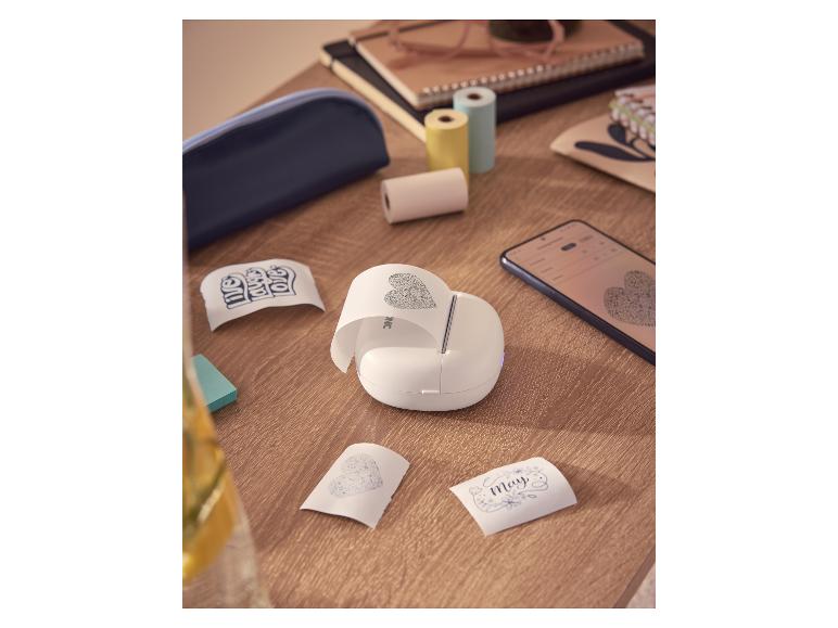 Imprimante portable blanche imprimant des autocollants en forme de cœur, sur une table en bois avec des rouleaux de papier.