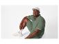Homme en polo vert, short et casquette, assis sur fond blanc.