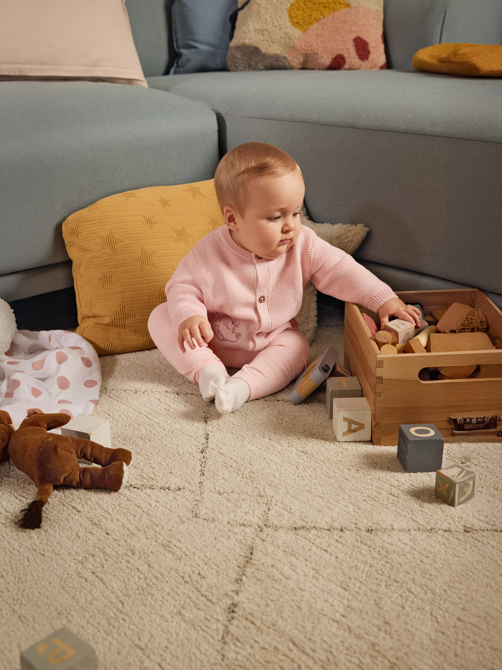 Bébé en grenouillère rose jouant avec des blocs de bois sur un tapis près d'un canapé.