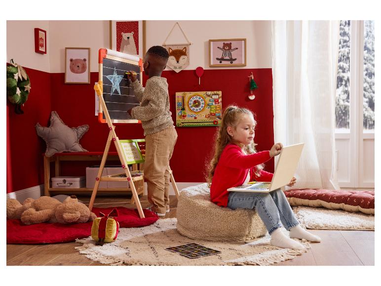 Deux enfants jouent dans une pièce décorée pour Noël : un garçon dessine sur un tableau, une fille utilise un ordinateur portable.