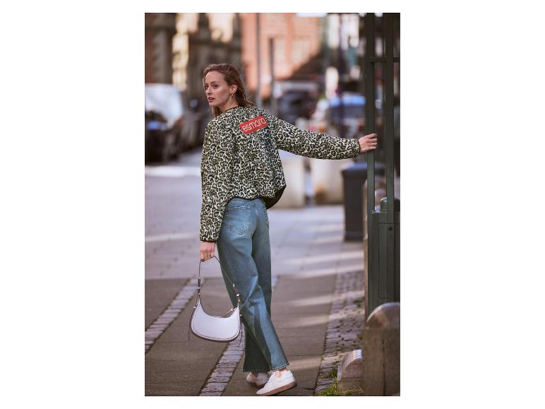 Une femme en veste léopard et jean tient un sac blanc dans une rue urbaine.