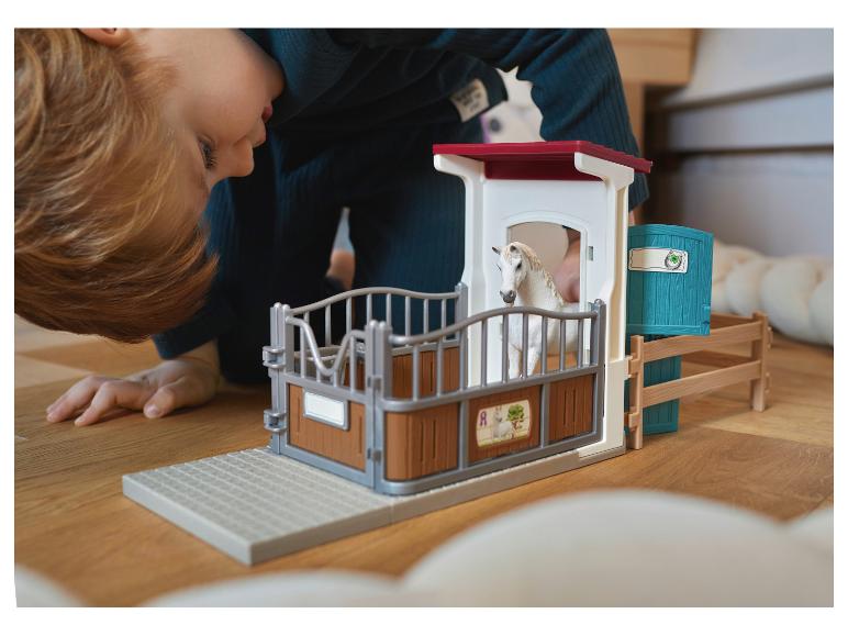 Enfant jouant avec une écurie jouet et un cheval blanc sur un plancher en bois.