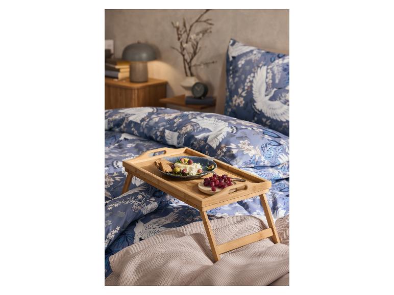 Un plateau de petit-déjeuner en bambou avec une assiette de nourriture et des raisins sur un lit avec de la literie bleu et blanc avec des grues.