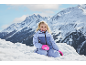 Fille en combinaison de ski et gants, riant dans la neige avec des montagnes en arrière-plan.