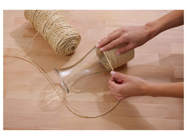 Mains enroulant une ficelle de jute autour d'un vase en verre sur une table en bois.