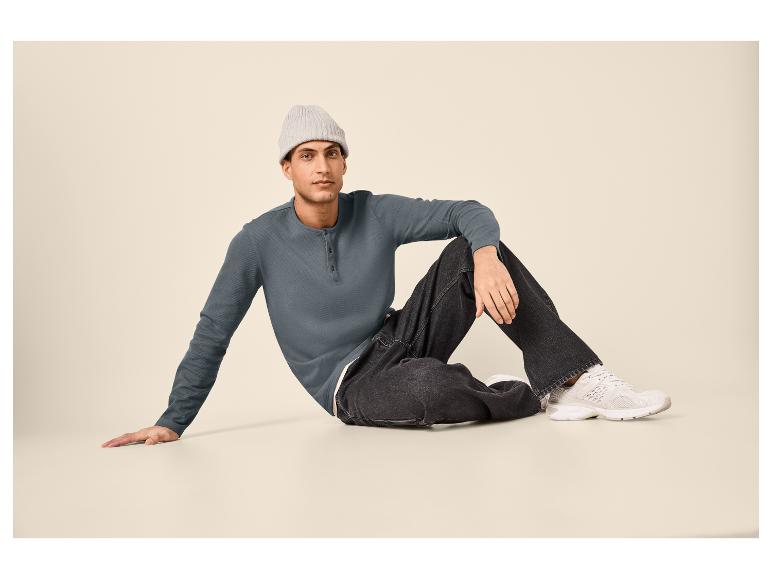 Homme en chemise henley grise, jean noir et baskets blanches assis par terre.