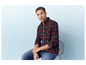 Homme en chemise à carreaux rouge et noire et jean bleu, assis sur une chaise.