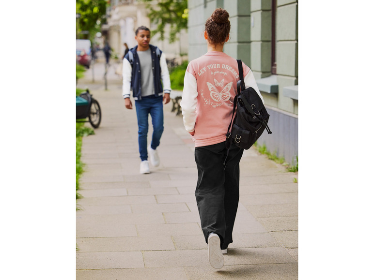 Une femme marche dans la rue avec une veste bomber rose et un sac à dos noir.