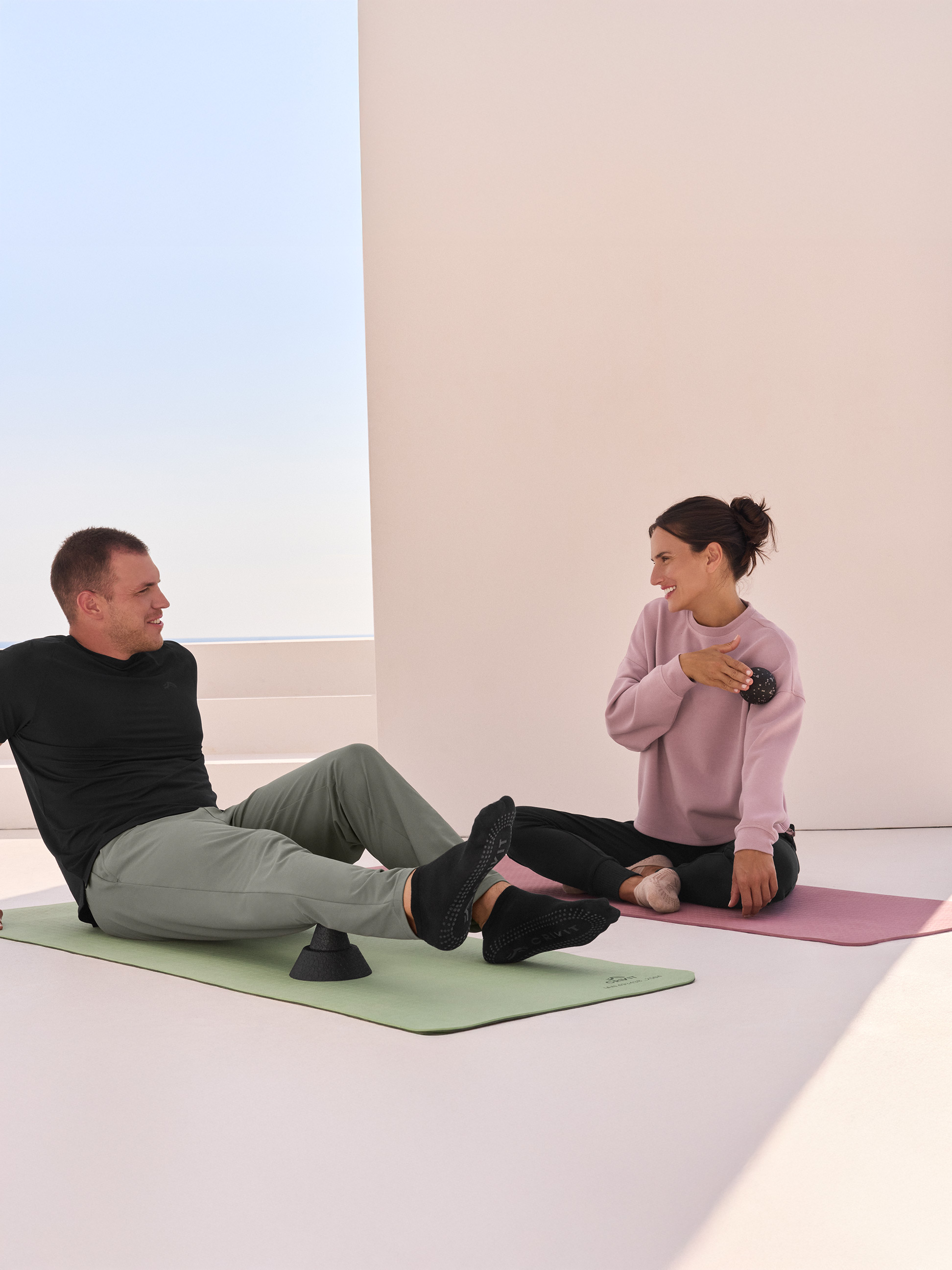Deux personnes sur des tapis de yoga, l'homme utilise un rouleau de massage, la femme une balle de massage.