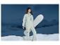 Femme en combinaison de ski verte tenant un snowboard devant les montagnes.