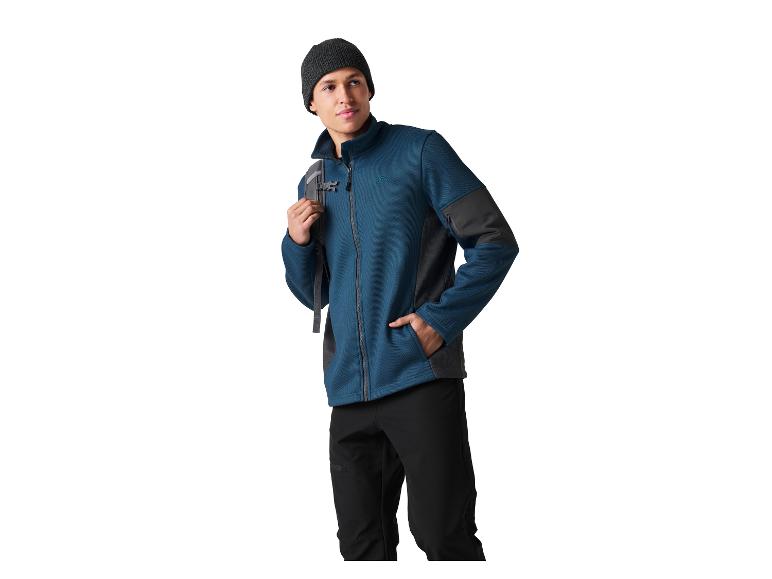 Homme en veste polaire bleue, pantalon noir et bonnet, avec un sac à dos sur l'épaule.