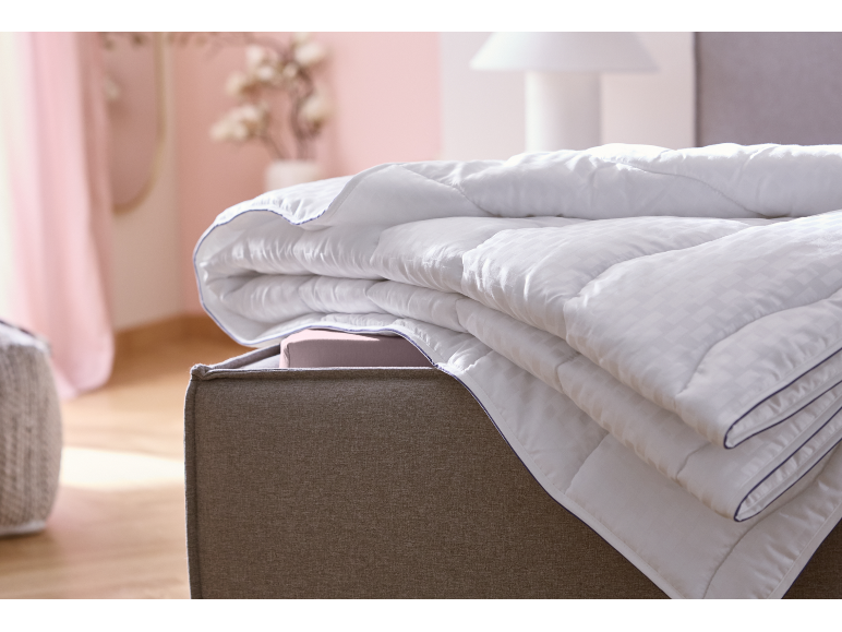 Couette blanche pliée avec liseré bleu sur un pouf gris dans une chambre.
