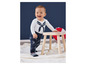 Un bébé dans un body blanc avec un nœud bleu et un pantalon bleu à motifs de bateaux se tient près d'un tabouret en bois.