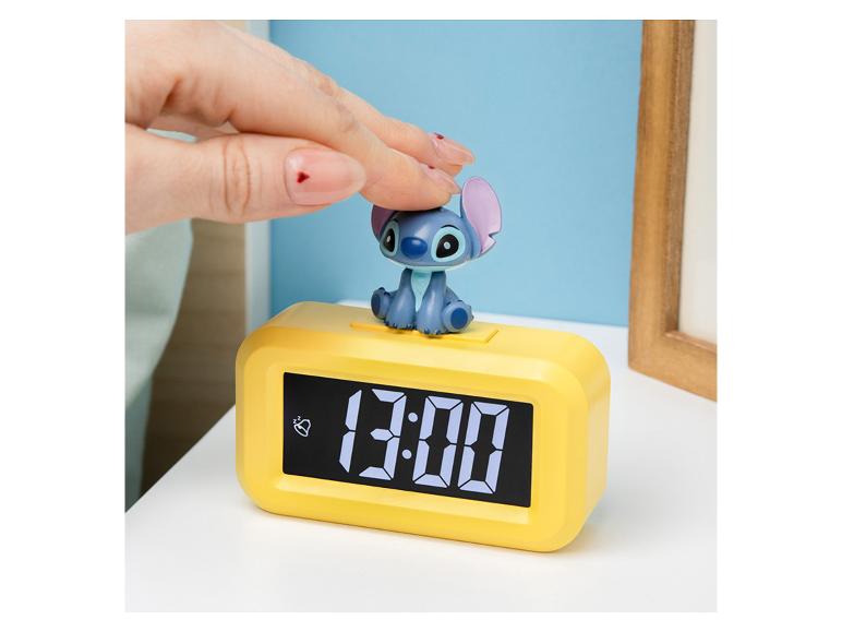 Une main appuie sur une figurine de Stitch posée sur un réveil numérique jaune affichant « 13:00 ».