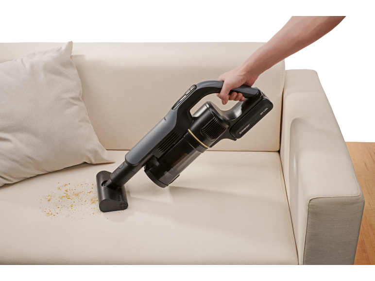 Aspirateur à main sans fil nettoyant des miettes sur un canapé beige.
