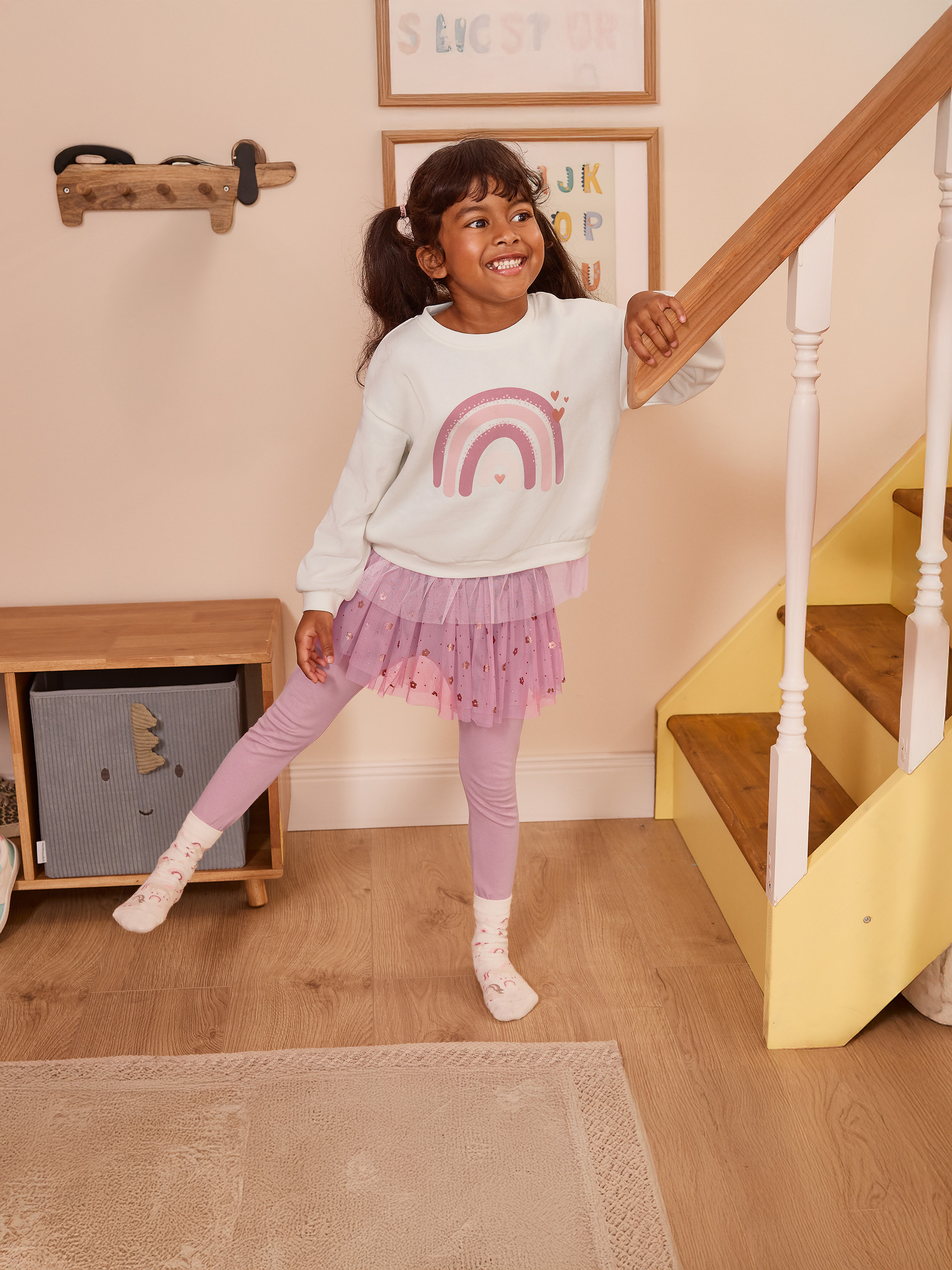 Fille en jupe en tulle rose et sweat-shirt arc-en-ciel, appuyée sur une rampe d'escalier.