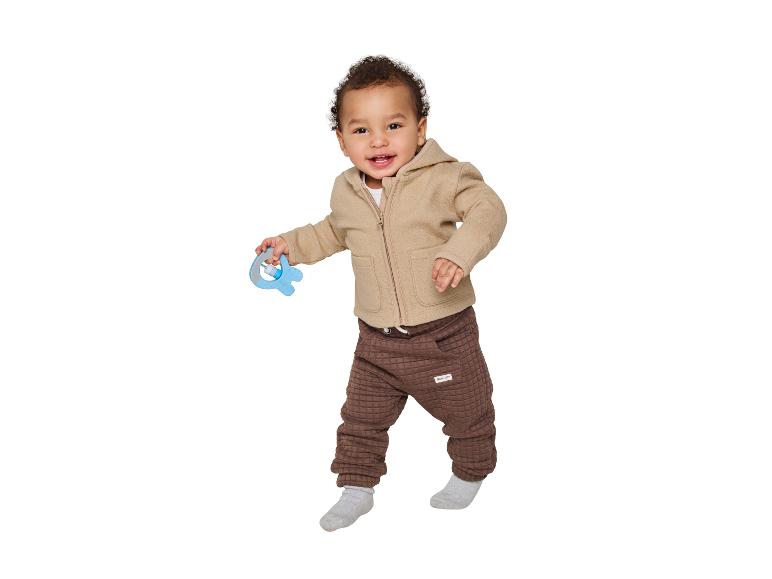 Bébé souriant portant une veste beige et un pantalon marron, tenant un hochet.