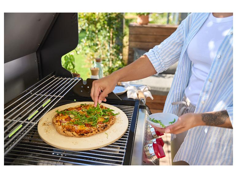 Une personne garnit une pizza de roquette sur un barbecue avec une pierre à pizza.
