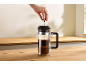 Une main presse le piston d'une cafetière à piston remplie de café sur un comptoir en bois.