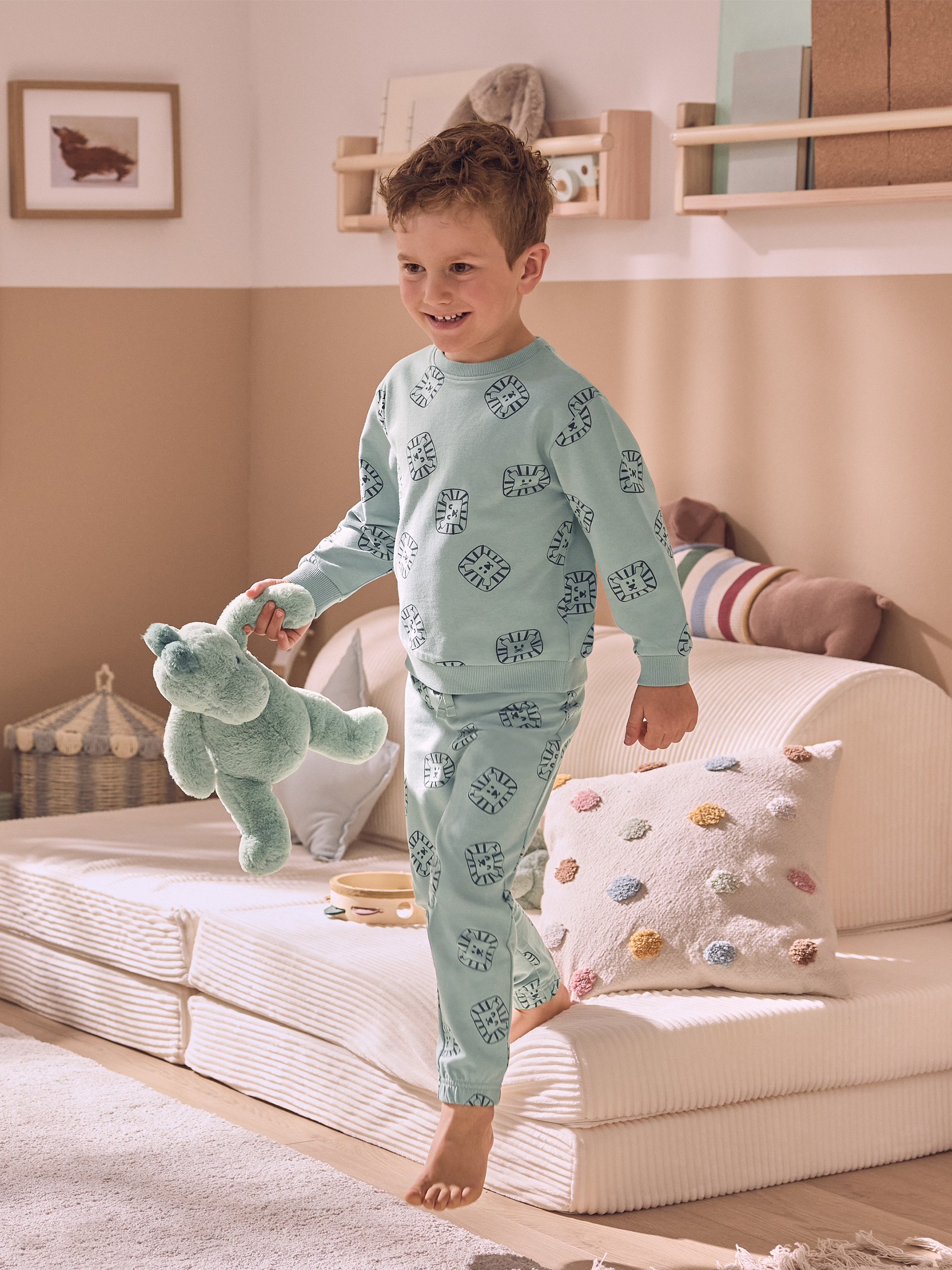 Un enfant souriant en pyjama bleu clair avec des motifs de lion, tenant une peluche verte.