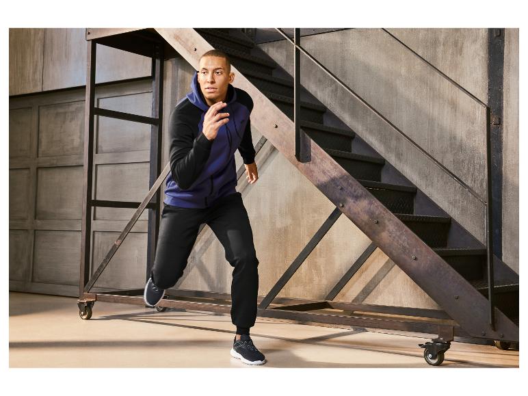 Un homme porte un sweat à capuche bleu et un pantalon noir dans un espace avec un escalier en acier.