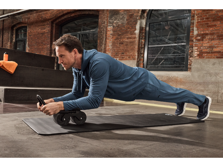 Un homme en survêtement bleu fait la planche sur un tapis de fitness noir avec une roue abdominale.