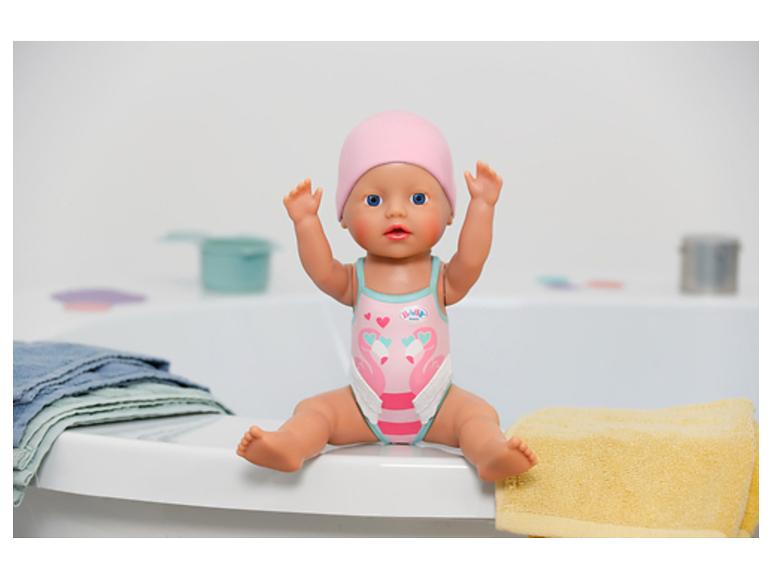 Poupée bébé en maillot de bain rose et bonnet, assise sur le bord d'une baignoire avec des serviettes.