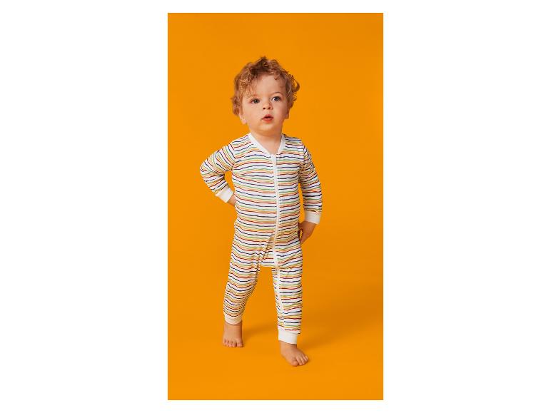 Un bébé aux cheveux bouclés en grenouillère rayée colorée debout sur un fond orange.