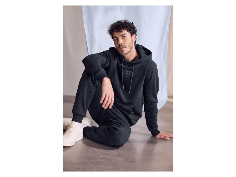 Homme aux cheveux bouclés foncés portant un ensemble sweat à capuche et pantalon de jogging noirs, assis.