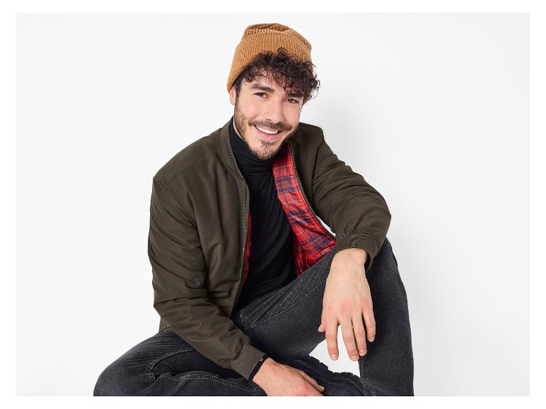 Homme avec bonnet marron, veste verte et col roulé noir, assis et souriant.