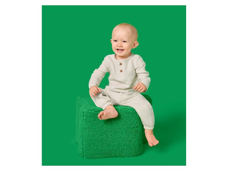Bébé souriant en ensemble de mousseline beige, assis sur un pouf vert.