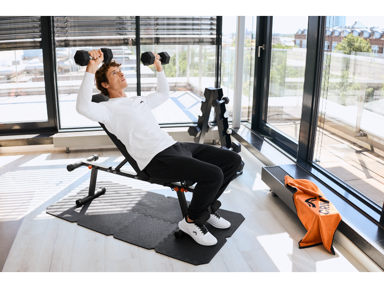 Homme soulevant des haltères sur un banc de musculation réglable dans une salle de sport à domicile avec un support à haltères.