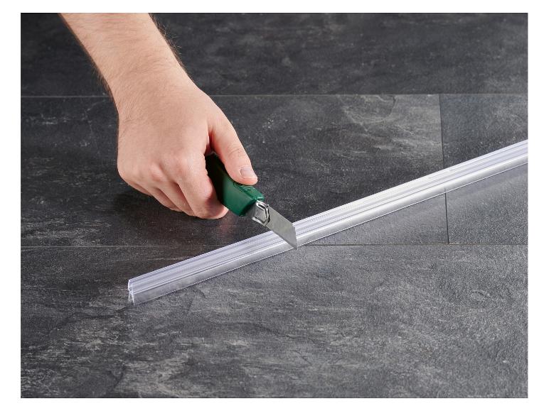 Main coupant un joint de douche transparent avec un cutter sur un sol carrelé.