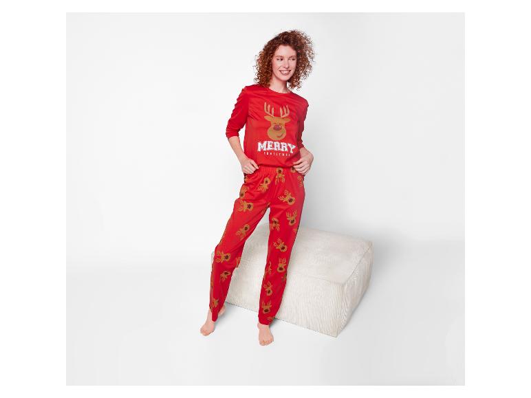 Femme aux cheveux roux bouclés portant un pyjama de Noël rouge avec des rennes, debout près d'un pouf.