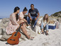 Un groupe d'amis se détendent sur la plage avec une guitare, un chien et une glacière.