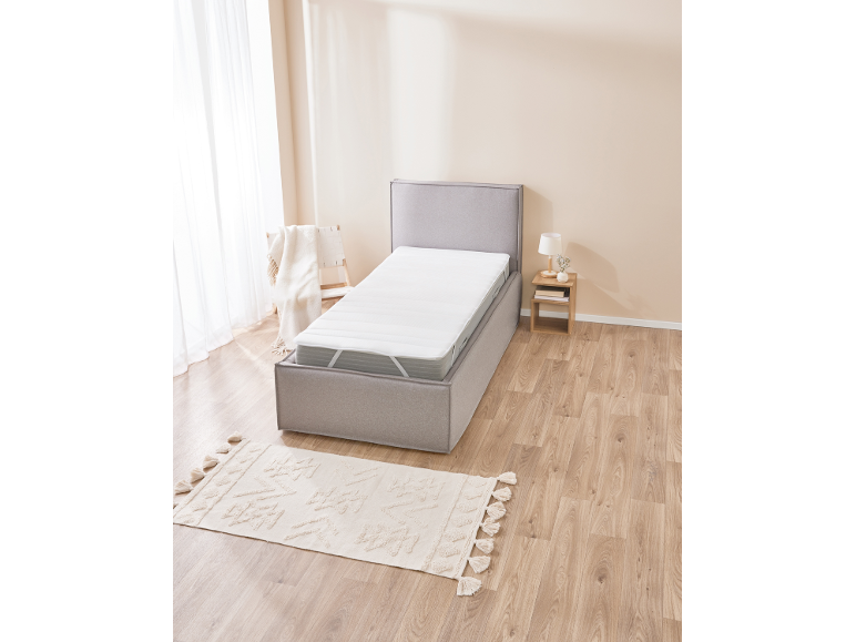 Lit simple avec revêtement gris, matelas blanc et tapis sur un parquet en bois.