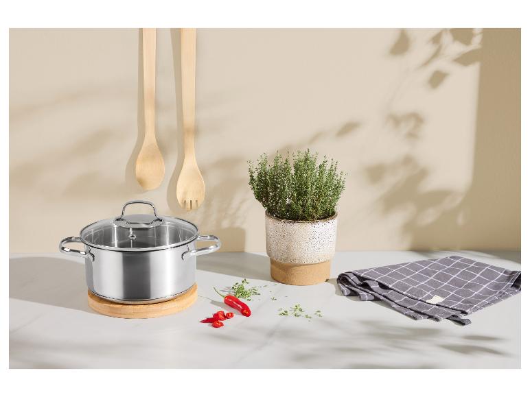 Casserole en acier inoxydable avec couvercle en verre sur dessous de plat en bois, ustensiles et herbes.