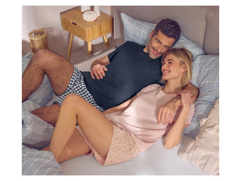 Couple en pyjama au lit, l'homme en t-shirt bleu et short à carreaux, la femme en t-shirt rose et short à motifs.