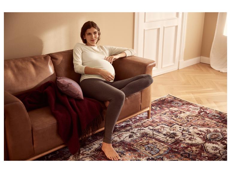 Une femme enceinte vêtue d'un haut blanc à manches longues et d'un legging gris est assise sur un canapé en cuir marron.