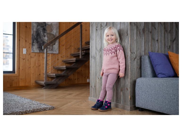 Fille souriante en pull et pantalon roses, avec des bottes, appuyée contre un mur en bois.