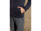 Un homme porte un pull bleu marine avec une fermeture éclair rouge et un pantalon gris.
